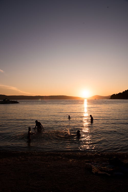 Silhouettes of unrecognizable travelers swimming in rippling sea with sandy beach against picturesque cloudless sunset sky