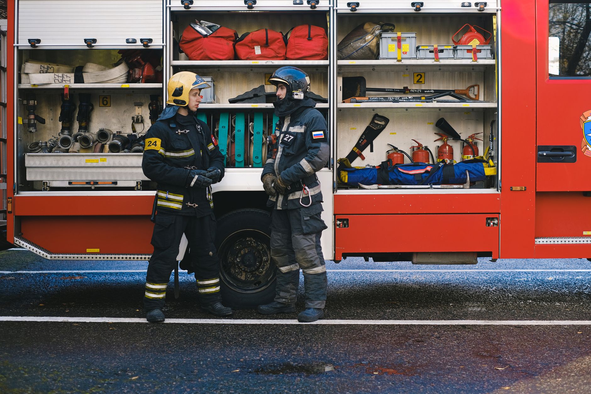 Two firefighters in full gear stand by a fire truck discussing strategy. Open equipment compartments visible.