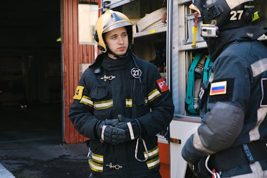 Two firefighters in uniform engage in discussion beside a fire truck ready for action.