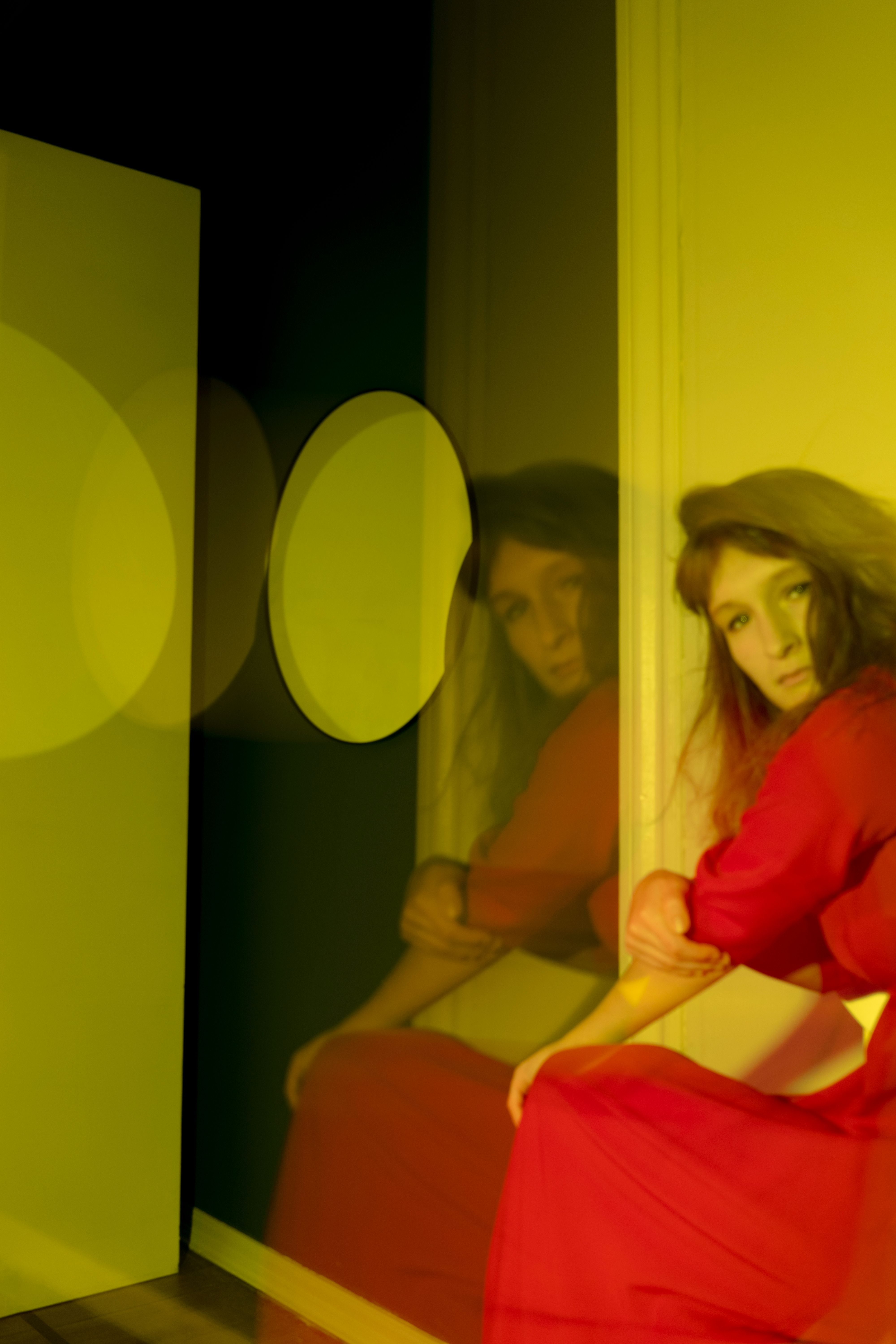 Blurred emotionless female in red maxi dress sitting in defocused room in yellow light and looking at camera
