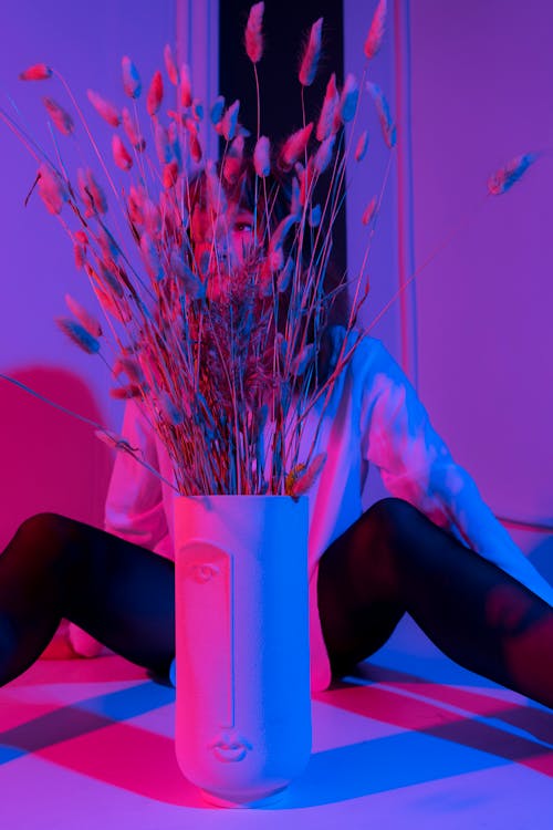 Emotionless female in black tights and white shirt sitting on floor with spread legs behind vase in dark room in pink and blue lights