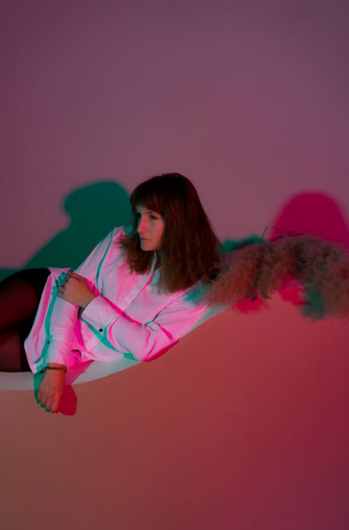 Contemplative adult female in tights and white shirt lying on curvy stand in dark room in pink lights and looking away