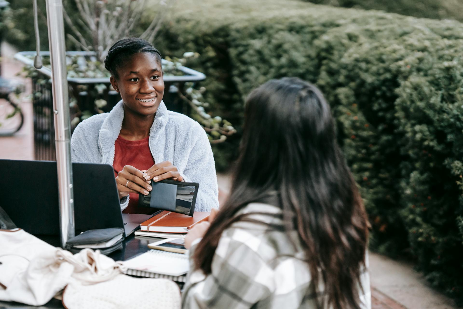 Positive multiethnic young female students sitting at table in campus with notebooks and laptop and talking about new project looking at each other