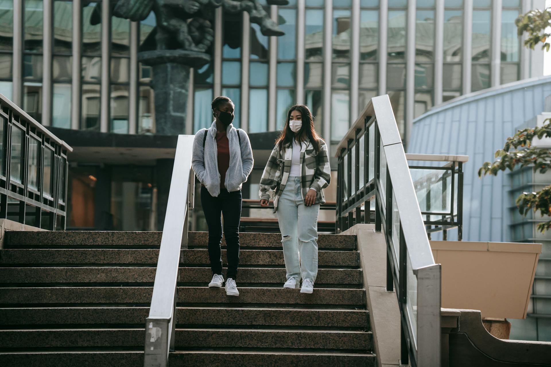 Full body young multiracial female students in casual outfits and protective masks going down stairs and talking in university campus