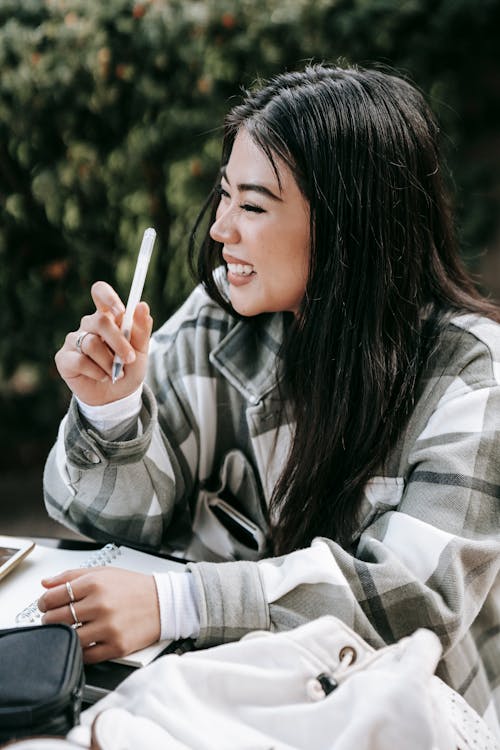 Free From above of cheerful young Asian female student in checkered shirt laughing while taking notes working on home task Stock Photo