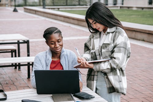 Free Cheerful young diverse female students with laptop taking notes while working together on research in university campus Stock Photo