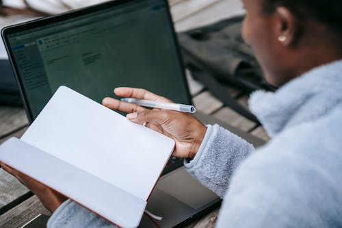 High angle of crop unrecognizable African American female student taking notes in notebook while working on research using laptop in campus