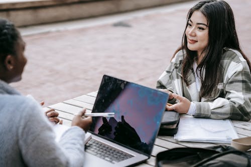 High angle of smiling diverse female students using laptop and taking notes while doing research together at table in campus