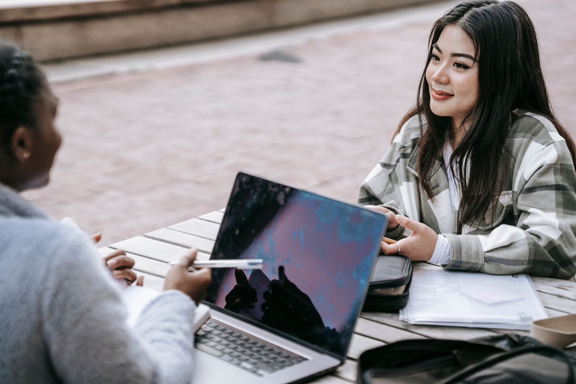 High angle of smiling diverse female students using laptop and taking notes while doing research together at table in campus