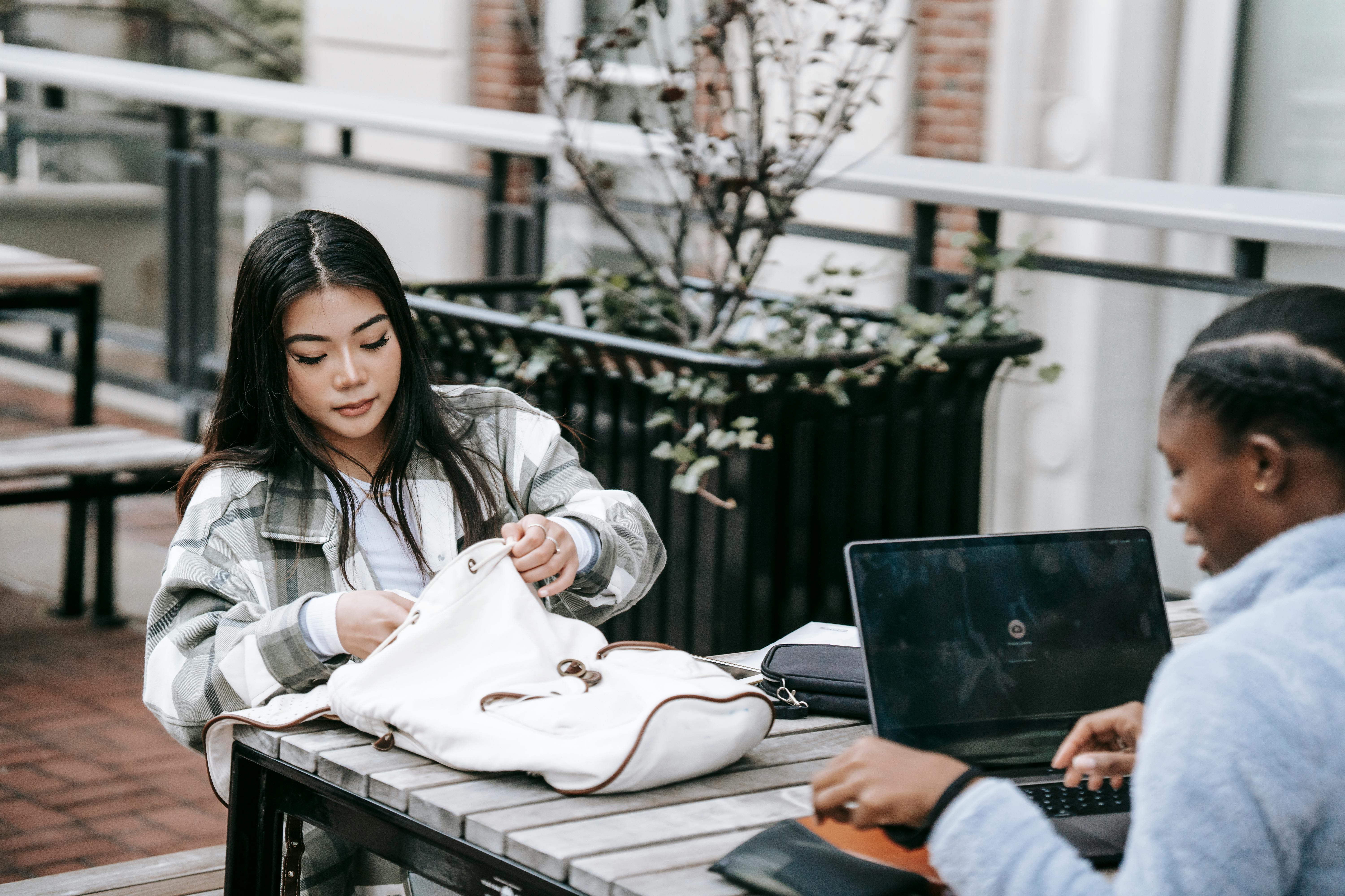 Young multiethnic female students sitting at wooden table in university campus while talking and working on project together using laptop