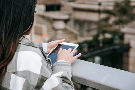 From above of crop unrecognizable young female in checkered shirt text messaging on tablet while standing near railing