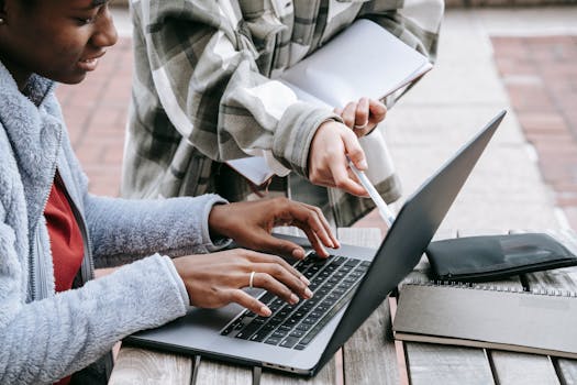 Two students collaborate on a laptop outdoors, focusing on a project together.