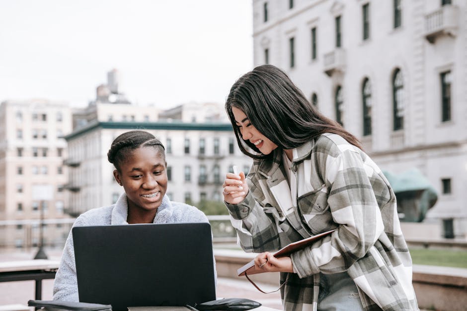 Young cheerful multiethnic female friends watching netbook while studying in town against buildings in daylight