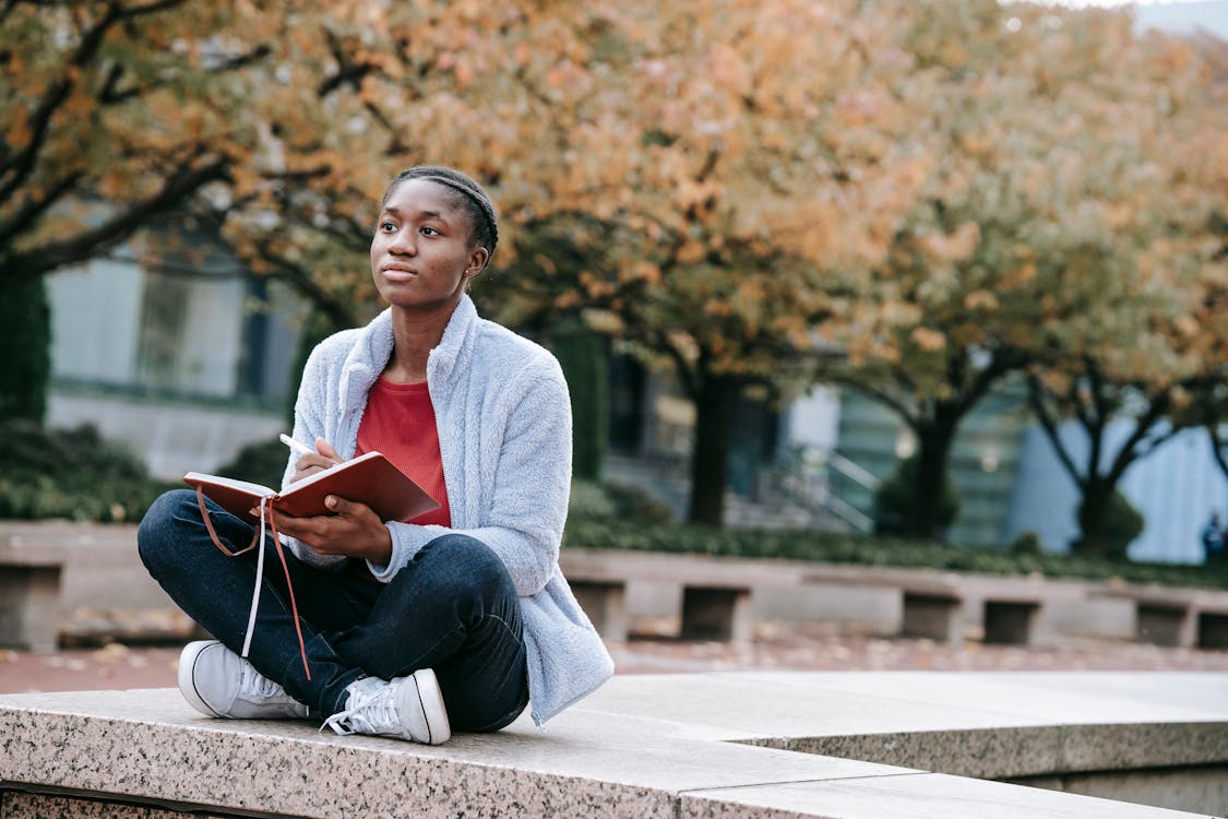 Free Young contemplative African American woman with agenda sitting with crossed legs against autumn trees while looking forward in city Stock Photo
