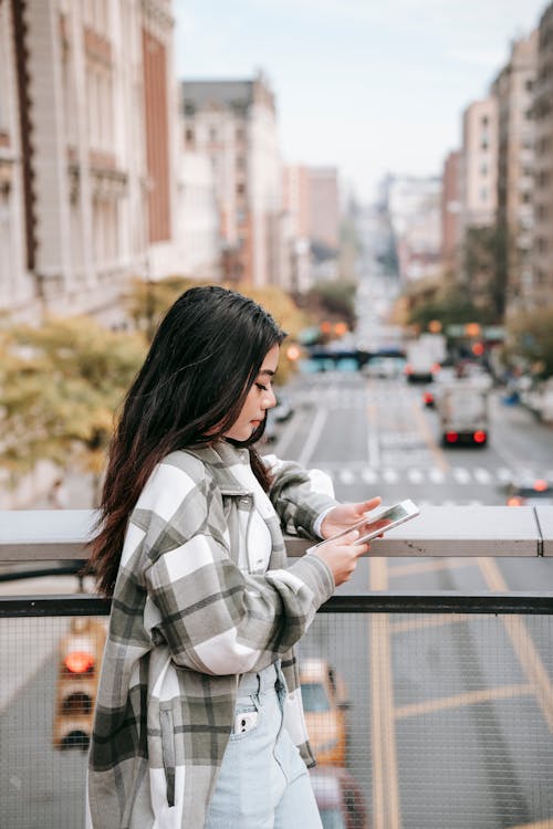 Side view of millennial ethnic female surfing internet on cellphone on bridge over city roadway in daylight