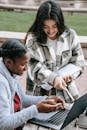 Cheerful diverse female students sharing laptop in city