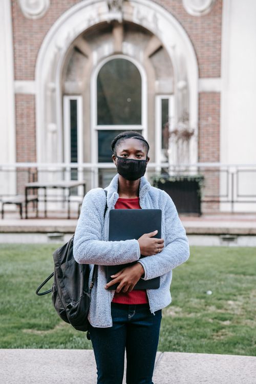 Free Anonymous ethnic female in mask with netbook and rucksack looking at camera against aged urban house during COVID 19 pandemic Stock Photo