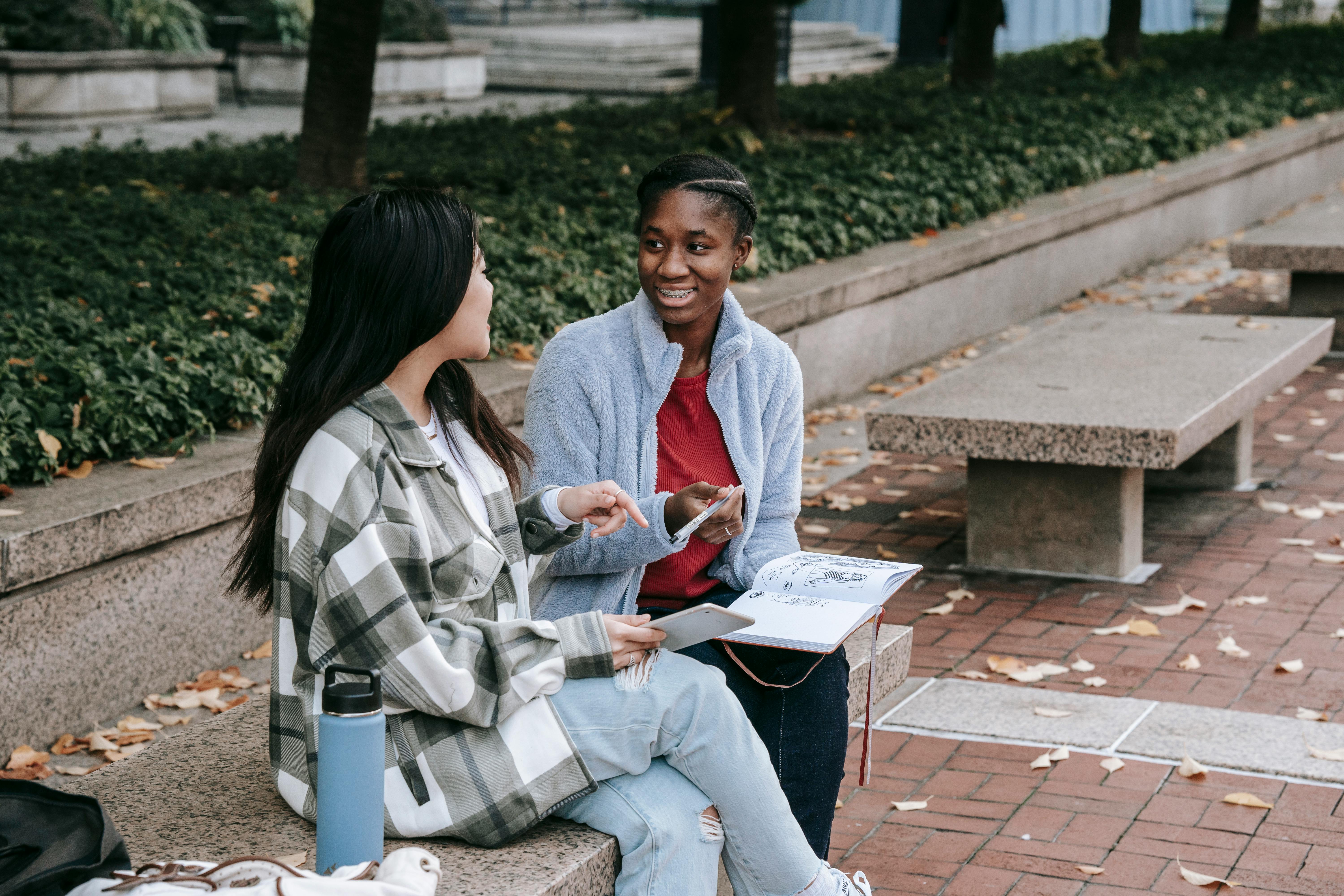 happy black woman talking to unrecognizable girlfriend on city bench