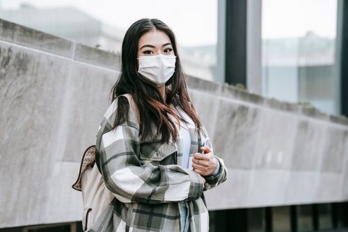 Free Young ethnic female in ornamental apparel and sterile mask with backpack looking at camera on city street Stock Photo