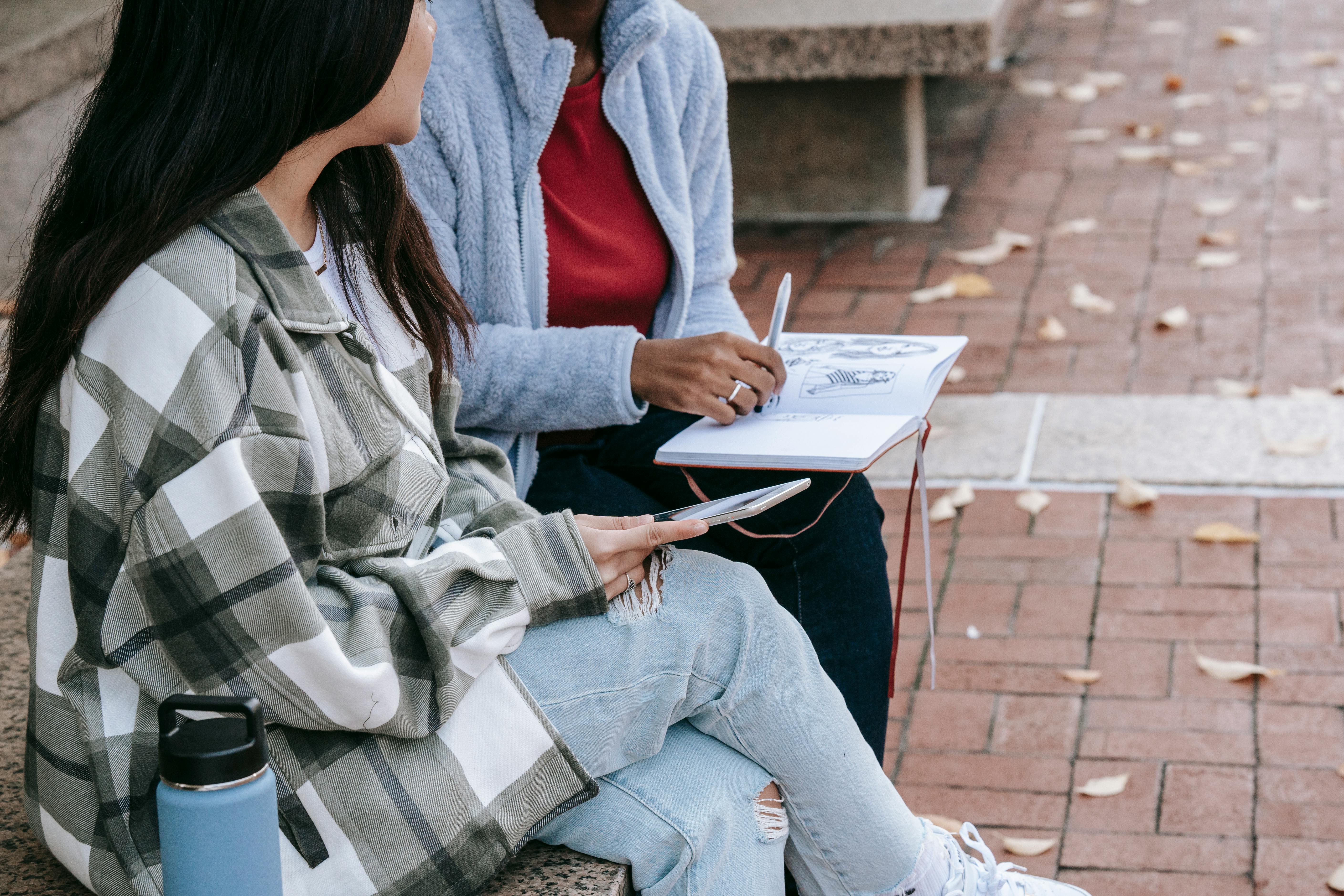 crop diverse girlfriends with diary and smartphone on urban bench