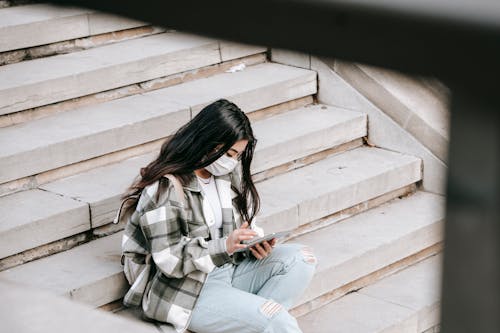 From above of anonymous concentrated young Asian lady with long dark hair in casual clothes and medical mask sitting on stairs on street and messaging on mobile phone