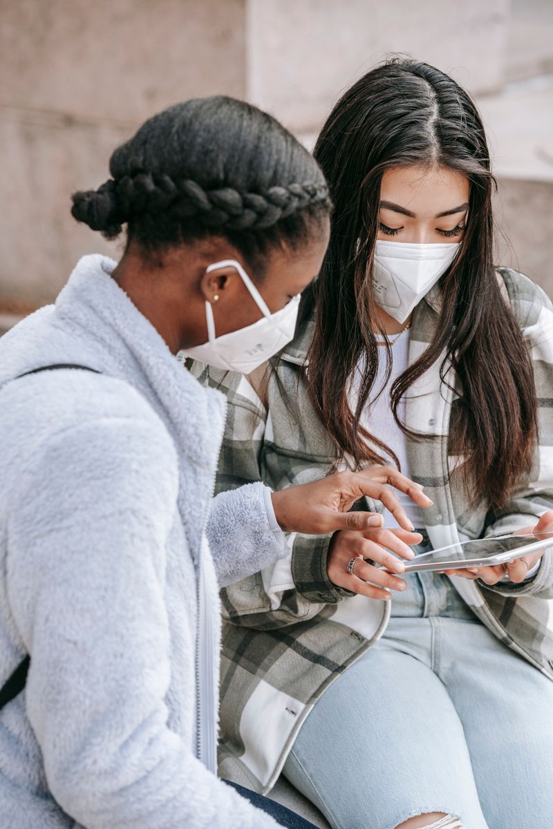 Anonymous multiethnic female teens in casual clothes and face masks sitting on bench and browsing tablet together after studies on sunny day