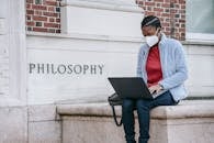 Concentrated young African American female student in casual clothes and medical mask sitting near Philosophy faculty and typing on laptop