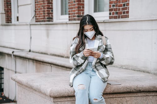 Free Young Asian female student with long dark hair in casual outfit and protective mask using smartphone near college building Stock Photo