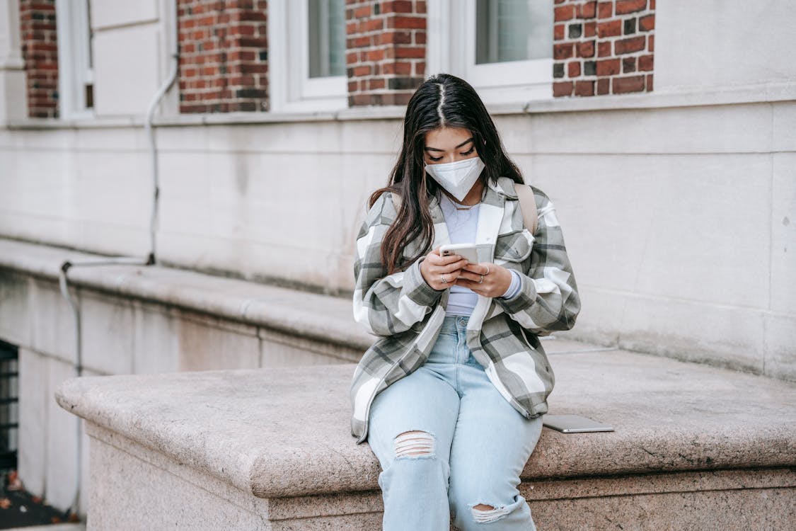 Young Asian female student with long dark hair in casual outfit and protective mask using smartphone near college building