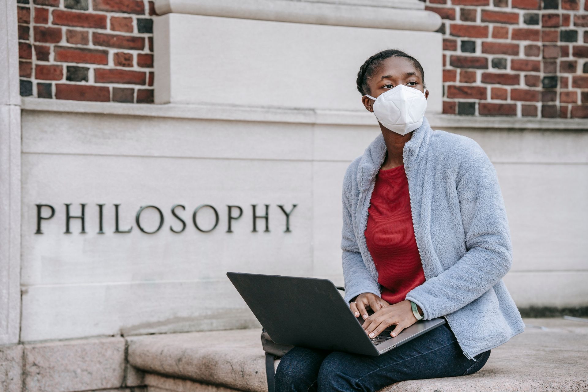 Young woman wearing mask, studying with laptop outside a philosophy building.