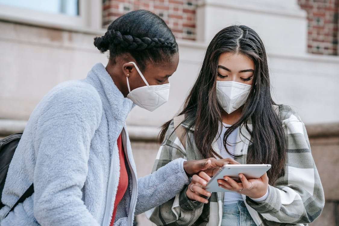 Focused young multiethnic female classmates in casual clothes and medical masks using tablet together while standing near brick building