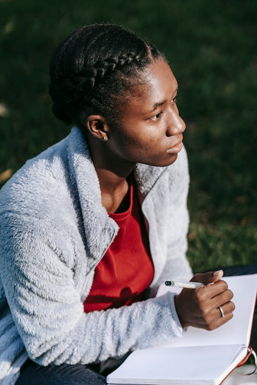 Side view of thoughtful young black lady in casual outfit sitting on grassy lawn while making notes notebook with pen in daytime