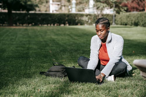 Full length of young African American lady in casual clothes with backpack sitting on grassy lawn while studying on computer in sunny day with legs crossed