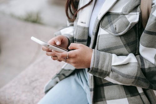 Crop anonymous young woman in casual outfit sitting on stone fence and text messaging on phone in daytime