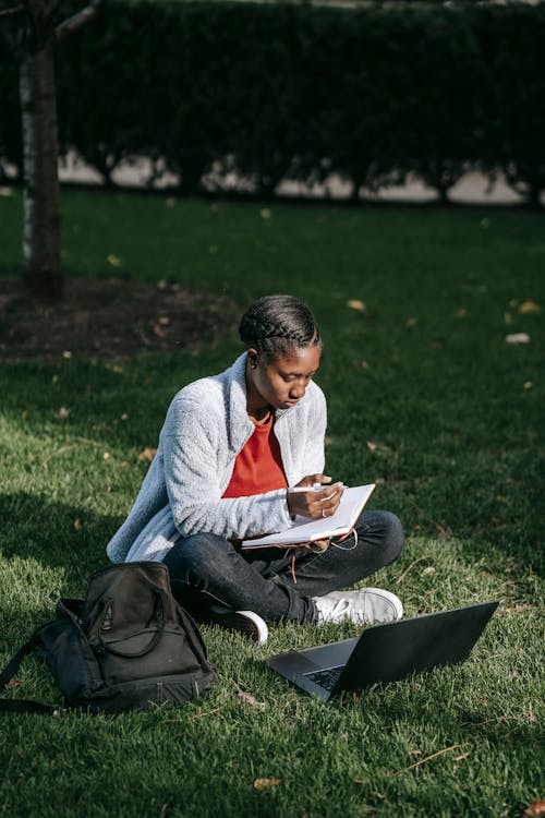 Full body of young black woman in casual outfit with backpack sitting on grass while studying on netbook and making notes in notebook with pen in sunny day