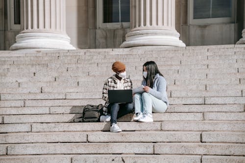 Free Full body of young multiracial female friends in casual clothes with protective masks sitting on stairs near building and using laptop while talking in daytime Stock Photo