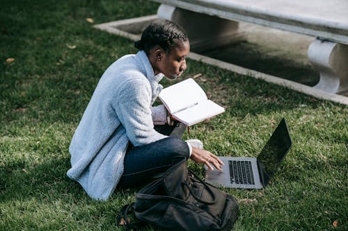 Side view of young concentrated African American female student working in project using laptop and taking notes while sitting on green lawn in campus
