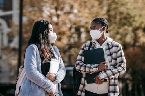 Young multiracial female students in casual outfit and protective masks with documents and laptop walking in university campus