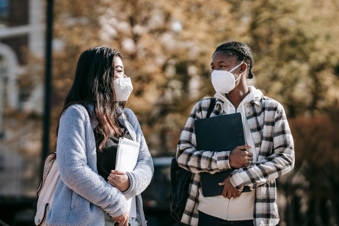 Free Young multiracial female students in casual outfit and protective masks with documents and laptop walking in university campus Stock Photo