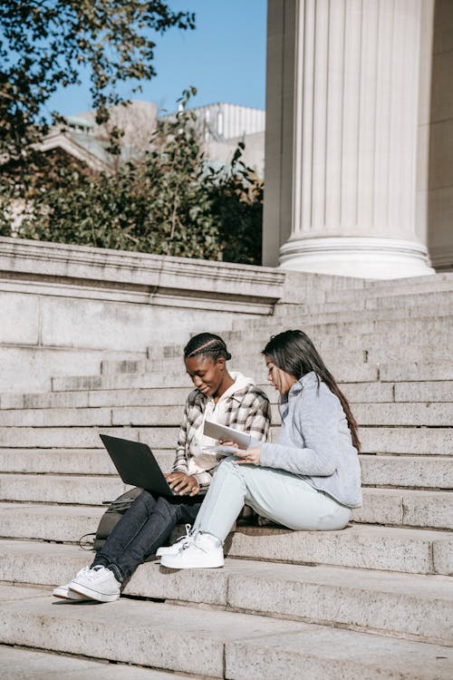 Full body young concentrated young diverse female students sitting on stairs in university campus and doing homework together using tablet and laptop