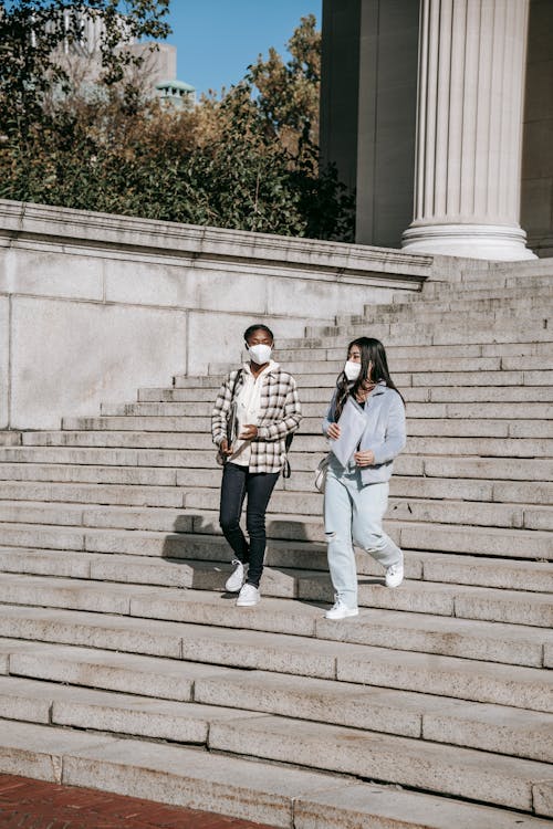 Full body young multiracial female students in protective masks and casual outfits with documents and laptop walking down stairs and talking in campus