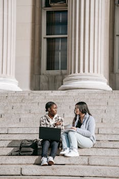 Full length young multiethnic female students talking about research working together while sitting on stairs in university campus