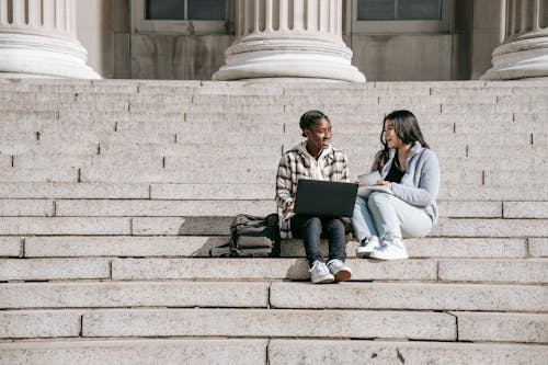 Full body positive multiracial female students sitting on stairs discussing research and working on laptop and tablet