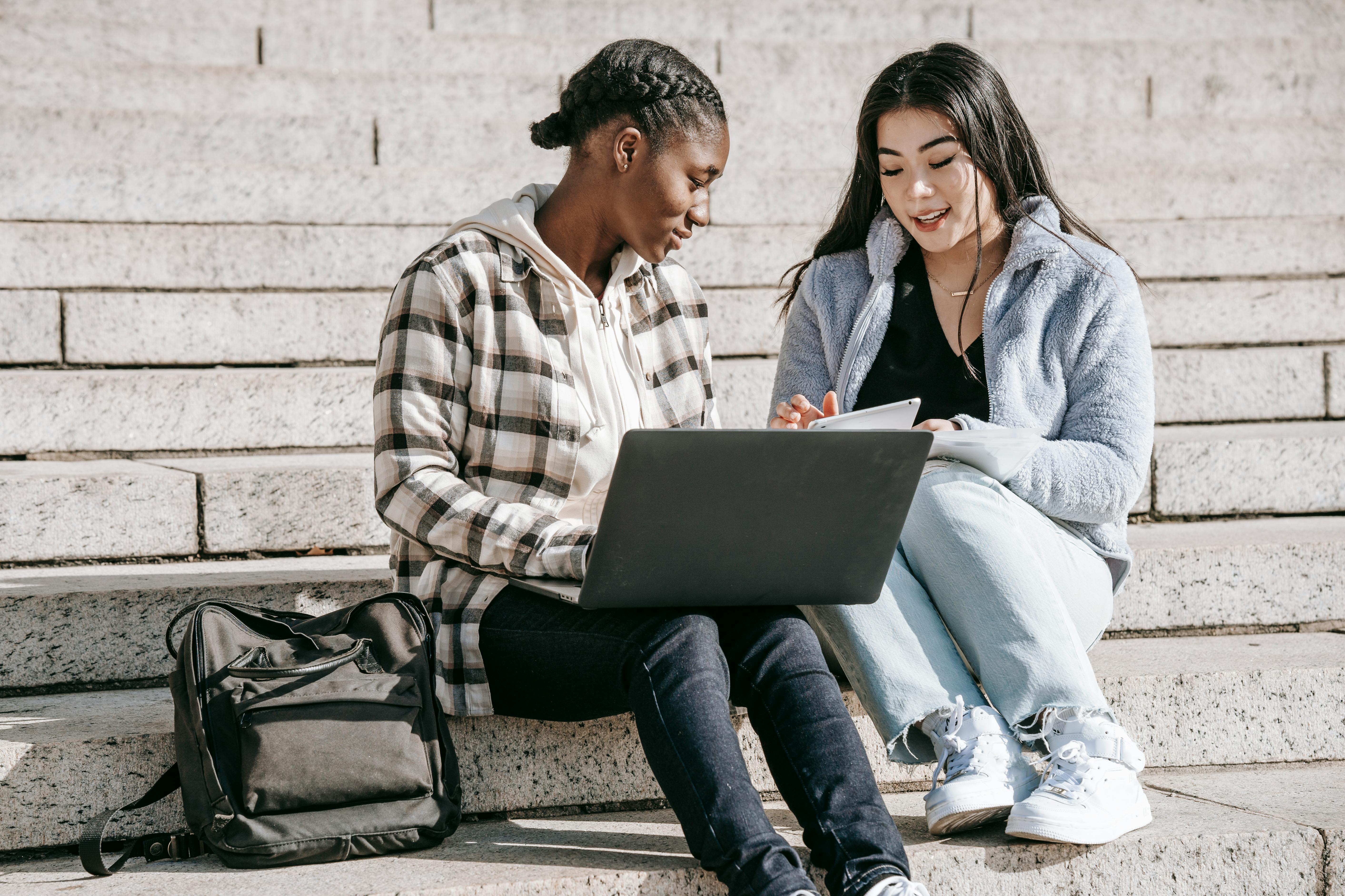 Two diverse students collaborating on laptops and notepads outdoors on university steps.
