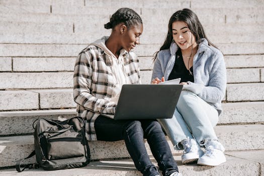 Two diverse students collaborating on laptops and notepads outdoors on university steps.