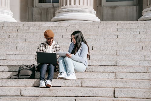 Full length cheerful young multiracial female students sitting on stairs with laptop and tablet while doing home task together
