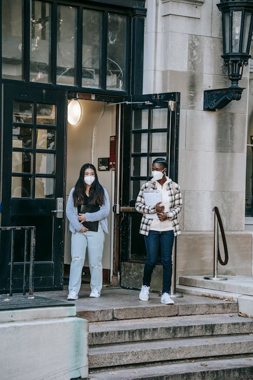 Full body multiracial female students in casual outfits and protective masks carrying folders and leaving college building together