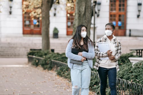 Calm young multiracial female students with folders wearing casual clothes and face masks strolling together on campus sidewalk