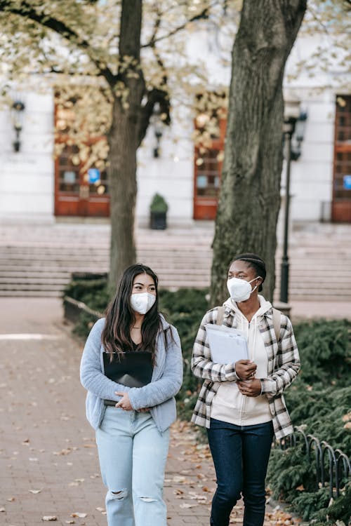 Young multiracial female students in respirators carrying folders and discussing plans while strolling together on campus sidewalk