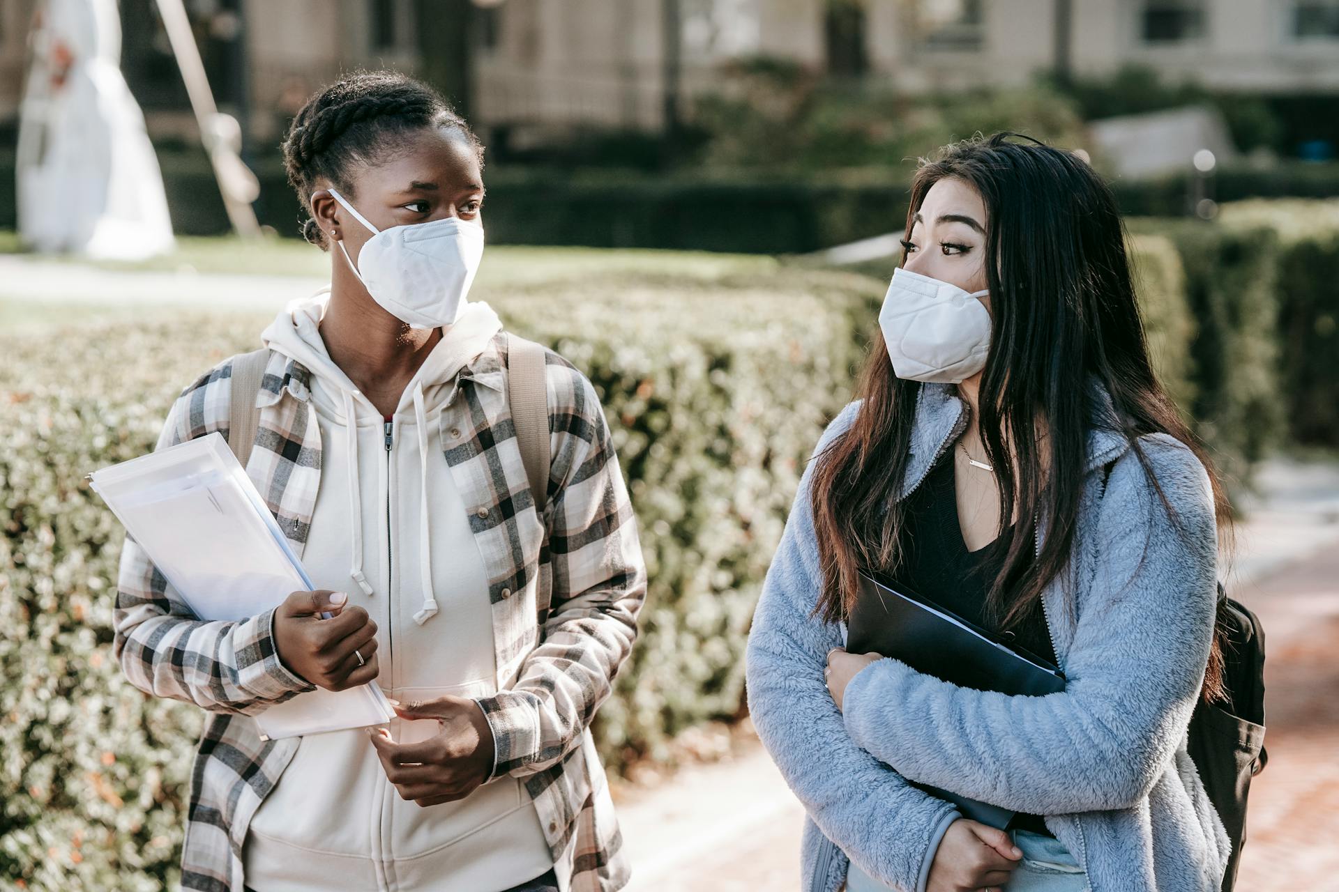 Positive young multiracial female students in protective masks carrying folders and chatting while standing on sidewalk on sunny day